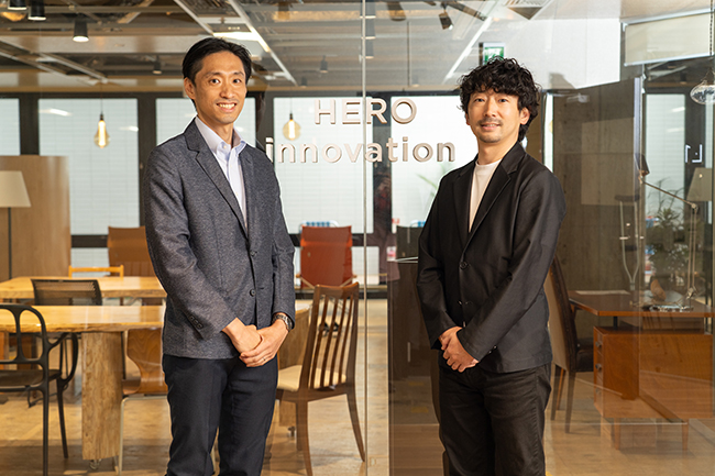 株式会社HERO innovationのロゴの前に立つ渡辺氏と井上講師